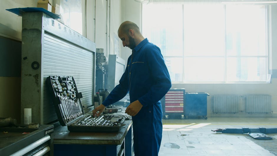 Mechanic inspecting tools in a workshop, wearing a blue jumpsuit with bright natural light from windows.